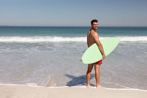 Smiling surfer standing at shoreline holding mint surfboard on sunny beach horizon