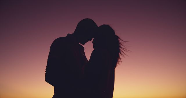 Silhouetted biracial couple shares a tender moment at dusk, with copy space. Their affectionate pose against the sunset creates a romantic outdoor setting.