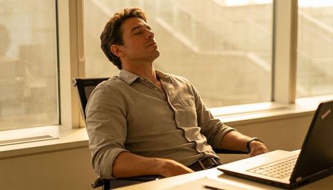 Calm office worker relaxing at desk, closing eyes by sunlit window with laptop in office