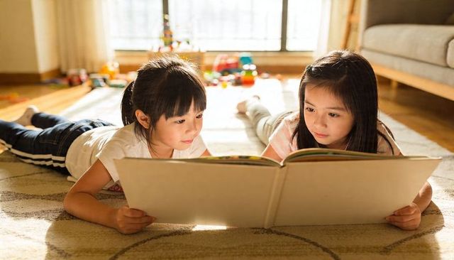 Two girls reading large picture book on sunlit living room rug sharing cozy moment