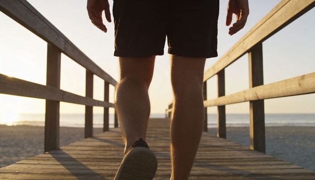 Walking toward sunset on wooden beach boardwalk showing male legs in shorts and sneaker by ocean