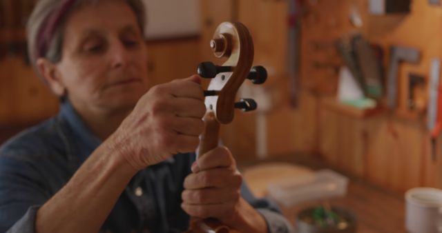 Front view close up of a senior Caucasian female luthier winding the strings on the tuning pegs on a finished violin in her workshop, inspecting the neck and scroll