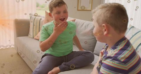 Two boys talking and laughing on living room sofa with patterned cushions, sunlit home