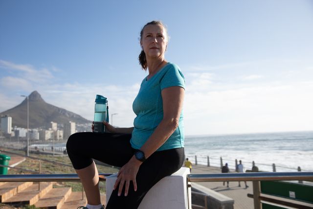 Senior Caucasian woman working out on promenade by the sea wearing sports clothes, sitting taking a break and holding water bottle. Retirement healthy lifestyle activity.