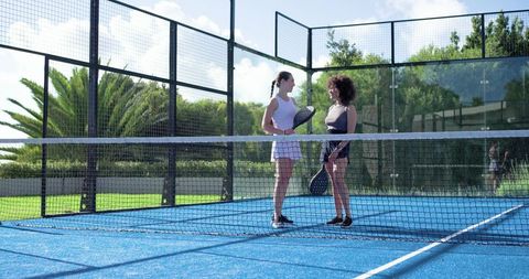 Diverse female padel players meeting at net on vibrant blue outdoor court under sunlight