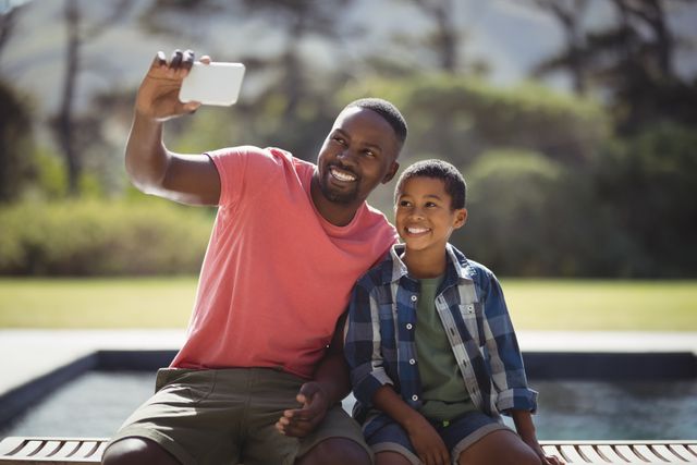 Father and son enjoying quality time together by the poolside, capturing a selfie with a mobile phone. Ideal for use in advertisements, family-oriented content, parenting blogs, and summer vacation promotions.