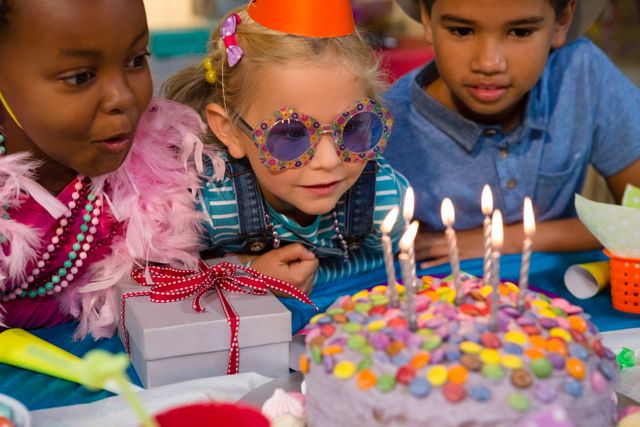 Children gathered around a birthday cake with lit candles, celebrating a birthday party. The scene is festive with colorful decorations, gifts, and joyful expressions. Ideal for use in advertisements, party planning materials, children's event promotions, and social media posts highlighting celebrations and joyful moments.