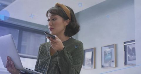 Asian businesswoman speaking into smartphone while holding laptop in modern office interior