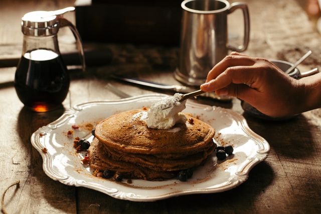 Hand adding whipped cream on stack of pancakes on rustic table. Surrounding items include syrup bottle, metal mug, knife, blueberries. Ideal for breakfast-themed articles, cooking blogs, and food magazine layouts.