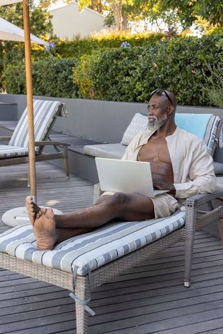 Senior african man relaxing on patio lounger using laptop for remote work and leisure