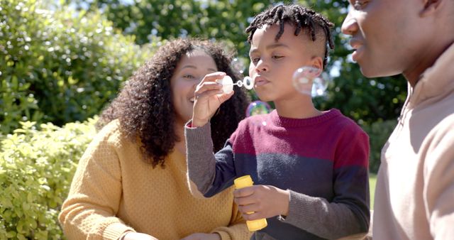 Child blowing bubbles with parents watching and smiling in garden during a sunny day. Ideal for depicting family bonding, outdoor activities, happiness, and leisure. Suitable for parenting blogs, family-oriented advertisements, and playful lifestyle promotions.