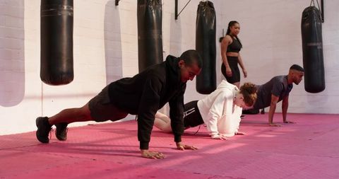 Diverse group doing push-ups in boxing gym on pink mat with heavy bags and trainer coaching