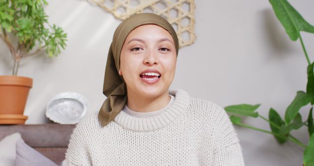 Image of happy biracial woman in hijab making image call talking to camera at home. Happiness, relaxation, communication, inclusivity and domestic life.