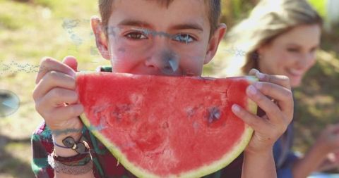 Young boy biting watermelon slice at sunny park wearing plaid shirt and bracelets