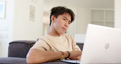 Young Asian man working from home on laptop, leaning on sofa and typing in modern livingroom