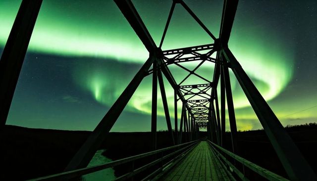 Green aurora over steel truss bridge at night with wooden walkway, leading perspective