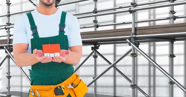 Builder dressed in overalls and tool belt holding a miniature house in hands while standing in front of scaffolding structure. Perfect for use in advertising real estate services, construction industry promotions, architectural design projects, and home renovation concepts.