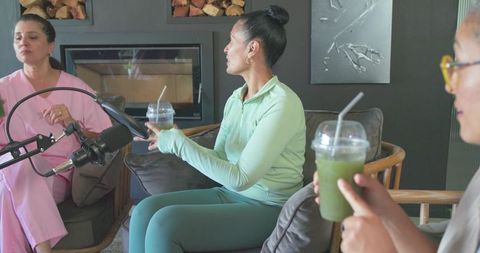 Women recording casual podcast in living room holding green smoothies by fireplace