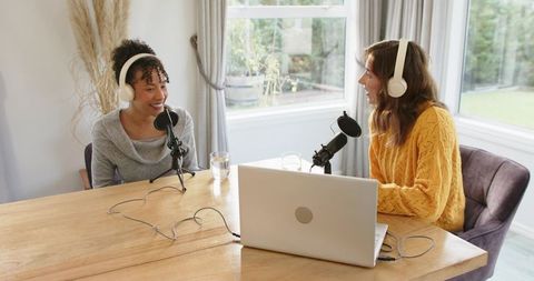 Two women recording cozy home podcast interview with laptop, microphones and headphones