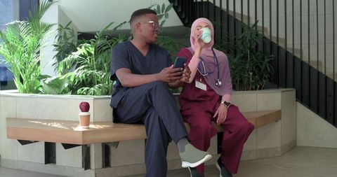 Diverse healthcare coworkers taking coffee break in bright atrium, woman in hijab sipping