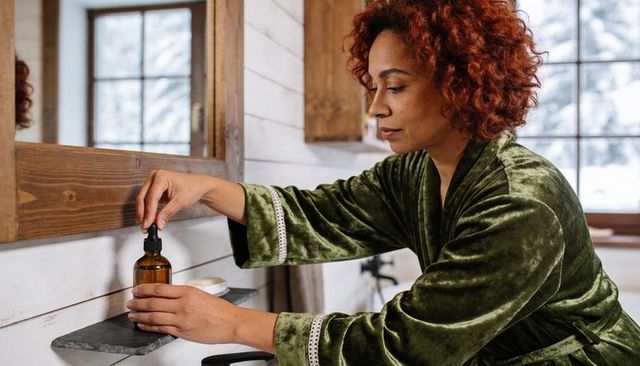 Woman in green velvet robe reaching for amber dropper bottle in rustic bathroom