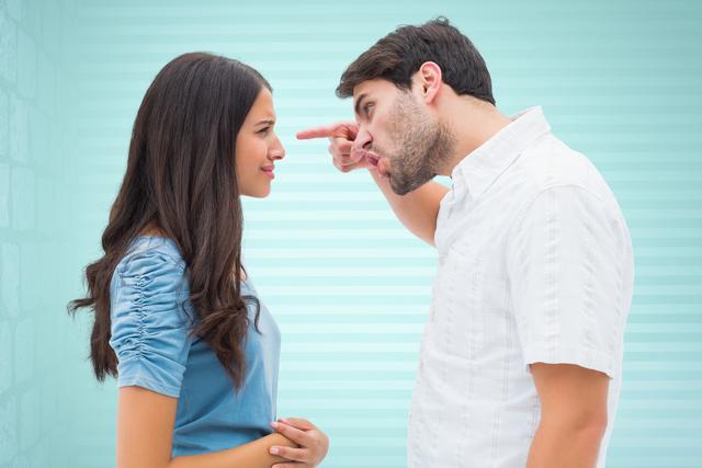 Young couple having disagreement indoors against a blue striped background. Man pointing and facing woman with intense expression, while woman looks composed. Suitable for illustrating concepts of conflict, communication issues, relationship dynamics, or emotional tension in romantic contexts.