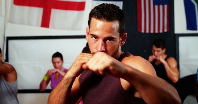 Group of individuals engaged in a boxing training session in a gym. A determined man in the forefront exercising punches wearing a maroon tank top. International flags in background suggest a diverse environment. Ideal for content related to fitness, boxing classes, motivation, and group workouts.