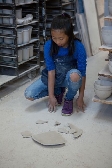 Girl in pottery workshop looking at broken plate pieces on floor. She is wearing a blue shirt and jeans with ripped knees. Shelves with pottery items are visible in the background. This image can be used to depict themes of learning, creativity, accidents, and the process of making ceramics. Suitable for educational materials, art class promotions, and articles about pottery and crafting.