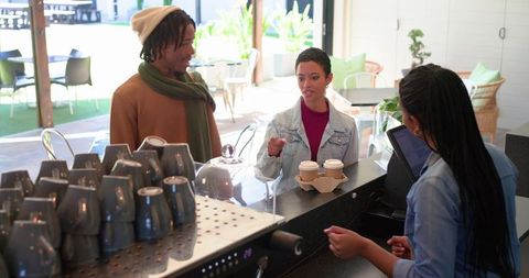 Young diverse friends ordering coffee at cafe counter while barista serves takeaway cups