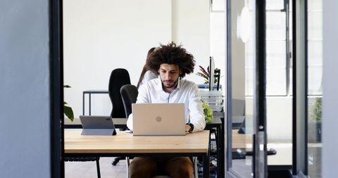 Young professional working on laptop in modern open-plan office with glass and plants