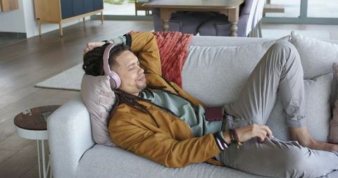 Relaxing mid-adult man listening to music on sofa wearing pink headphones and casual jacket
