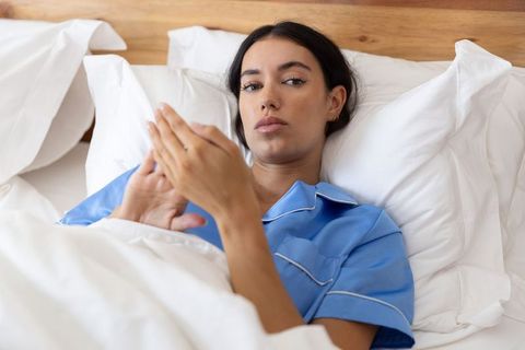 Young woman lying in white bed wearing blue pajamas using smartphone for evening relaxation