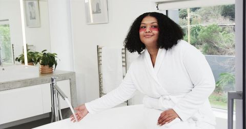 Relaxing self-care moment in luxury bathroom sitting on bathtub rim wearing robe and pink eye patche