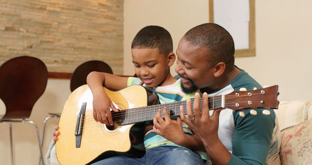 Father and son enjoying a moment playing guitar together at home. Ideal for topics around family bonding, parent-child relationships, music education, home activities, and parenting resources. Suitable for articles, blog posts, and advertisements focusing on parenting, family life, and learning musical instruments.