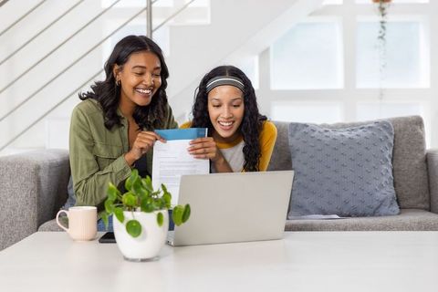 Mother and teen daughter opening mail together on living room couch with laptop