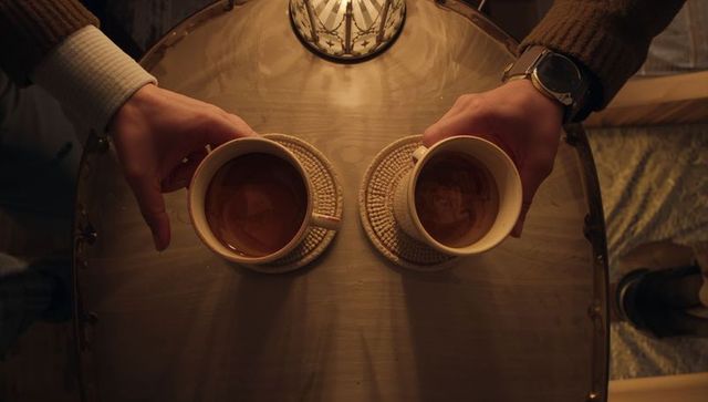 Hands holding two ceramic coffee cups on round table, cozy top-down view with warm lighting
