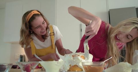 Friends laughing while spreading frosting on homemade layer cake in bright modern kitchen