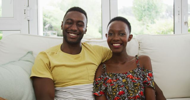 This image depicts a happy African American couple sitting on a sofa, embracing and smiling warmly at the camera. They appear content and relaxed, enjoying each other's company in a cozy home environment. This photo can be used in articles or advertisements that focus on relationships, domestic life, mental wellbeing during quarantine, or the importance of family and togetherness. It is also suitable for social media posts promoting positive lifestyle choices, home decoration, or inclusive representation in media.