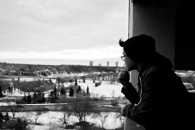 Black and white photo of man wearing glasses and beanie hat leaning on railing while gazing at expansive winter landscape featuring city buildings and bare trees, suitable for illustrating concepts of contemplation, solitude, urban life, and introspection in cold weather settings.