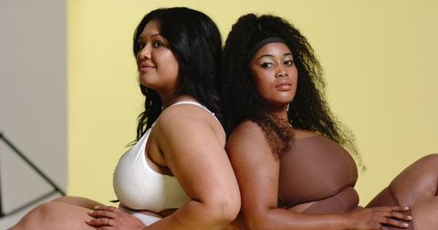 Curvy women posing back-to-back in swimwear on pastel backdrop, body-positive portrait