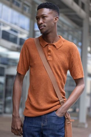 Young man wearing rust knit polo carrying brown crossbody bag standing in urban plaza