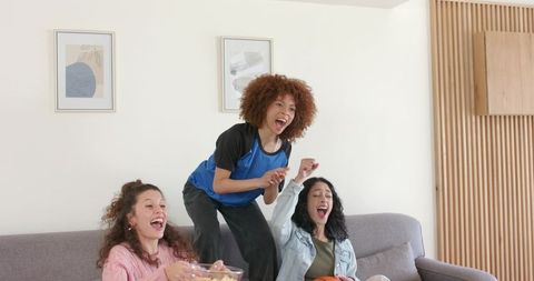 Three friends cheering on couch watching game with popcorn celebrating joyful home moment