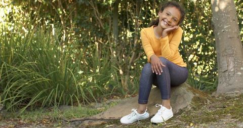 Smiling child sitting on mossy rock in sunlit woodland wearing yellow top and white sneakers