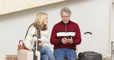 Senior couple waiting with luggage at terminal using smartphone and drinking coffee