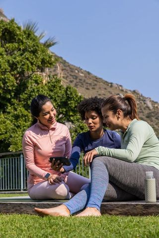 Multicultural women relaxing and sharing smartphone after outdoor workout in sunny park