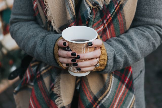 Woman wearing cozy plaid scarf holding hot beverage in disposable paper cup. Perfect for use in winter or autumn-themed promotions, outdoor clothing advertisements, content about comfort and warmth, or articles on seasonal fashion and lifestyle tips.