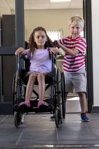 Boy pushing wheelchair with girl in school corridor, childhood inclusion and friendship
