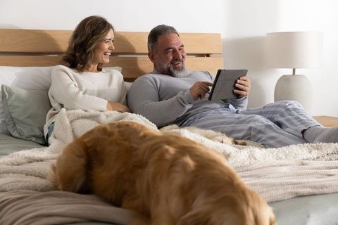 Mature couple relaxing in bed using tablet with golden retriever and cozy blankets
