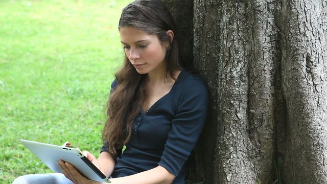 Young woman sitting under a tree in a grassy park using a tablet computer. The scene is serene and relaxed, with a focus on her casual use of technology in an outdoor setting. Suitable for themes relating to digital learning, leisure activities, nature, and technology. Ideal for promoting outdoor activities, digital education apps, and devices, or lifestyle articles.