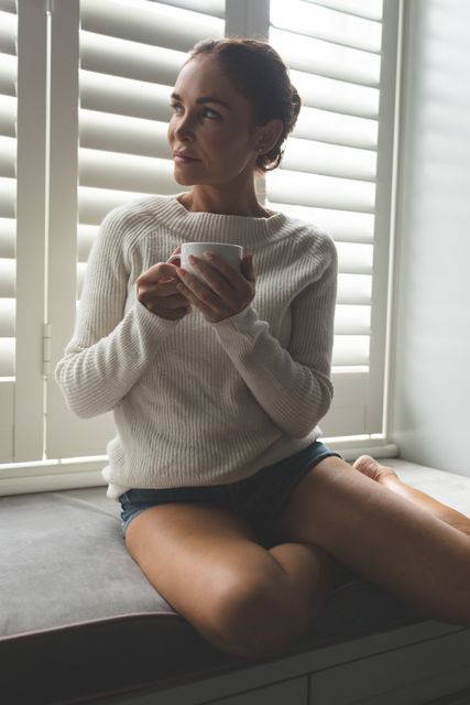 Front view of woman looking away while having a coffee on window seat in a comfortable home